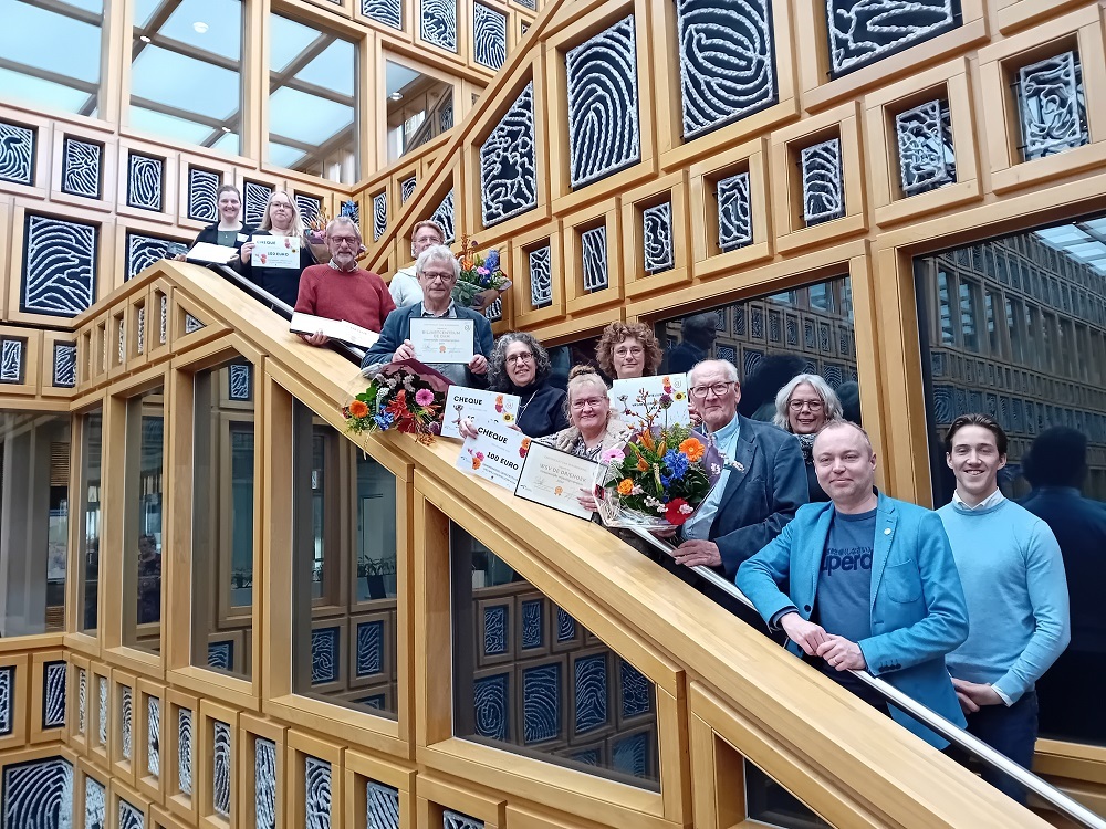 De winnaar van de Gemeentelijke Vrijwilligersprijs en de andere genomineerden staan met hun cheque opgesteld op een trap in het stadshuis, bij de vingerafdrukken die het gebouw typeren. Wethouder Rob de Geest is er ook bij.